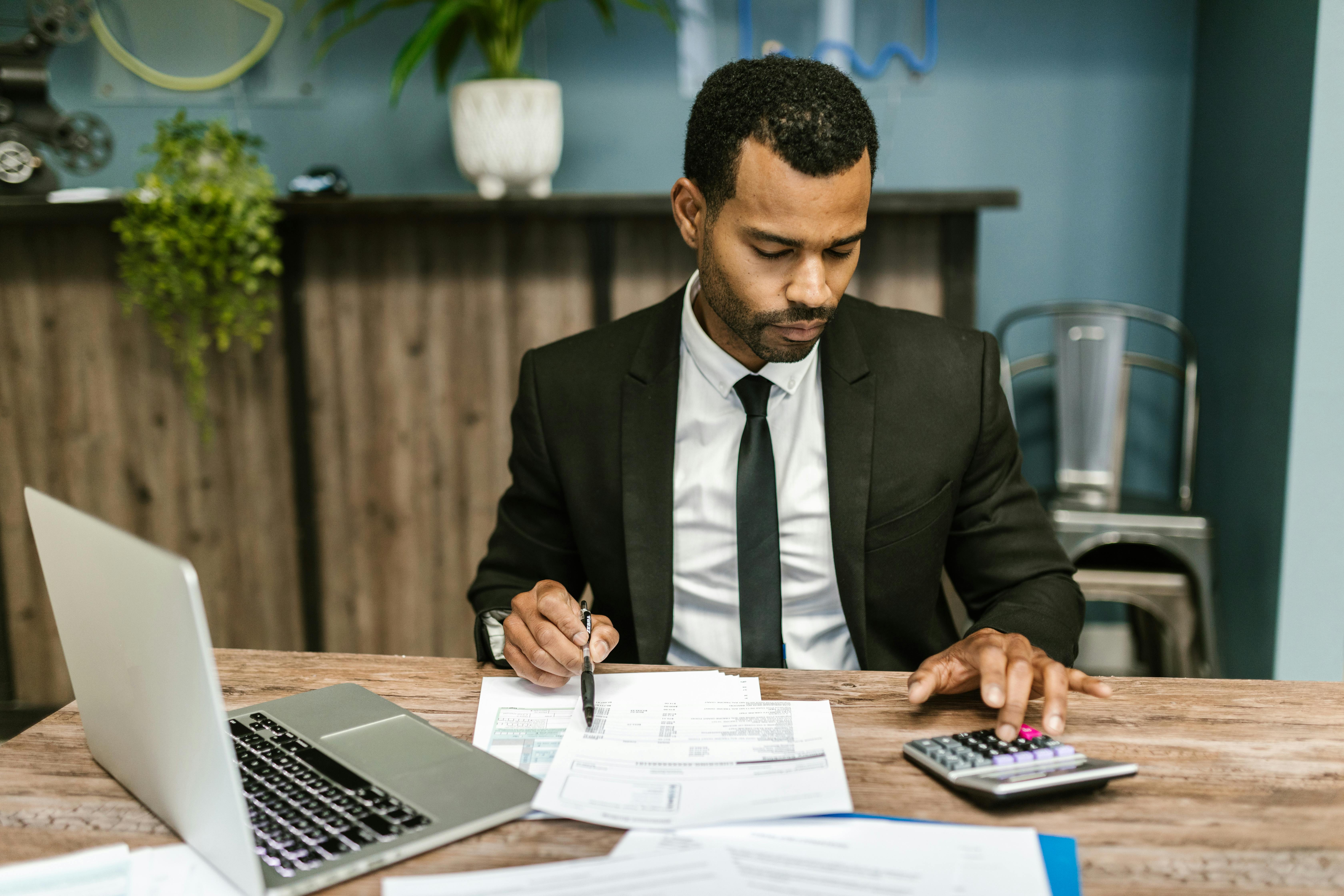 Man in black tie sitting at a desk with a laptop and calculator